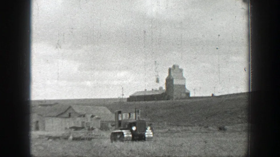 1937 California 8mm frame with a tracked tractor moving across a rural field below distant farm buildings and a tall agricultural structure.