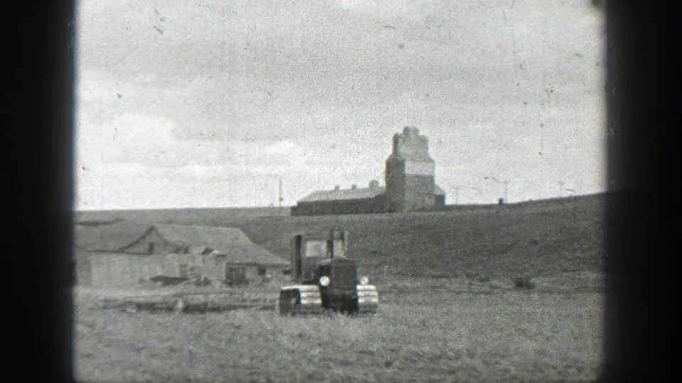 1937 California 8mm frame showing the tracked tractor near the center of open farmland, with low buildings and a tall farm structure in the background.