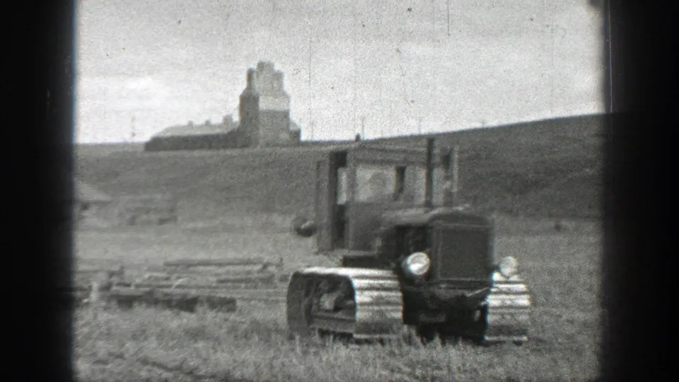 1937 California 8mm frame with a closer view of a small tracked tractor crossing rough field ground near distant agricultural buildings.