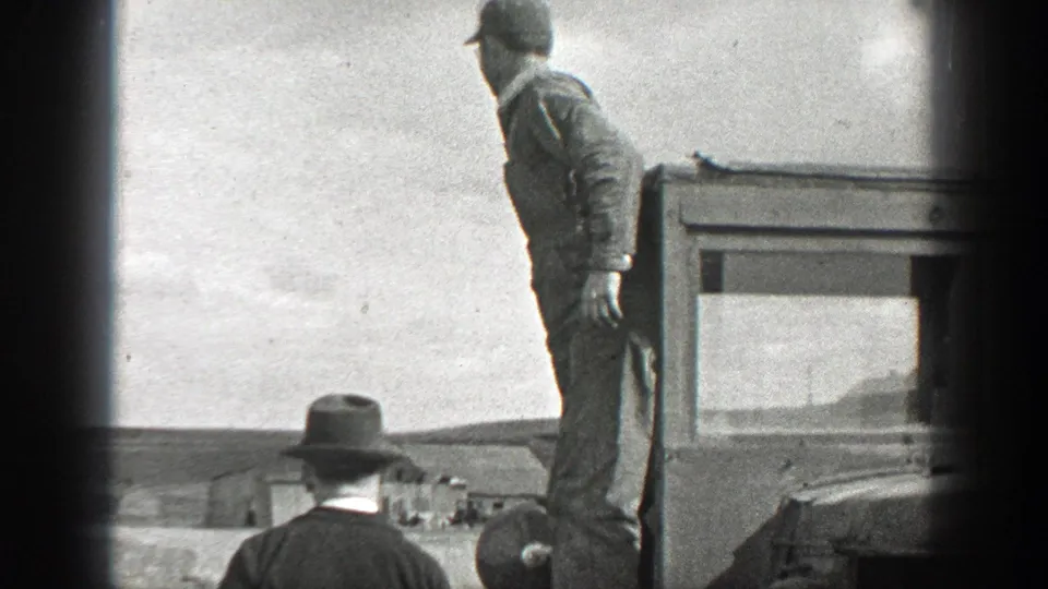 1937 California home-movie frame showing a man standing on a truck bed with another hat-wearing man below in an open outdoor setting.