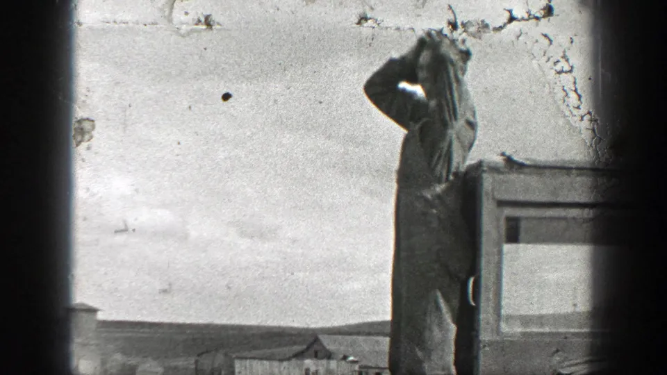 1937 California frame of a man in work clothes near a vehicle, lifting a hand to his hat or head against a bright open background.