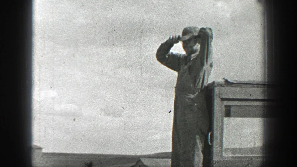 1937 California home-movie frame showing a worker in a cap shielding his eyes beside a vehicle, with open land behind him.