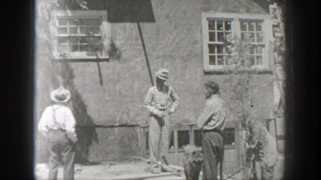 Three men stand near a stucco wall with multi-pane windows as a small dark dog moves at their feet.