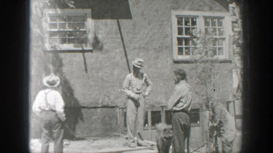 1937 California home-movie frame showing three men by a stucco building with windows while a small dark dog stands near them.