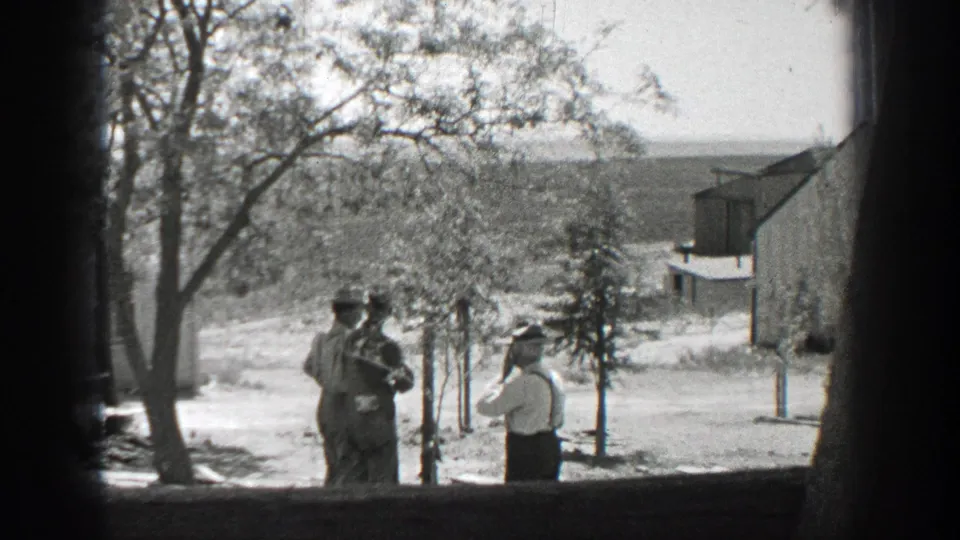 1937 California frame showing men gathered under a tree near fencing, with a distant rural building and hillside visible.