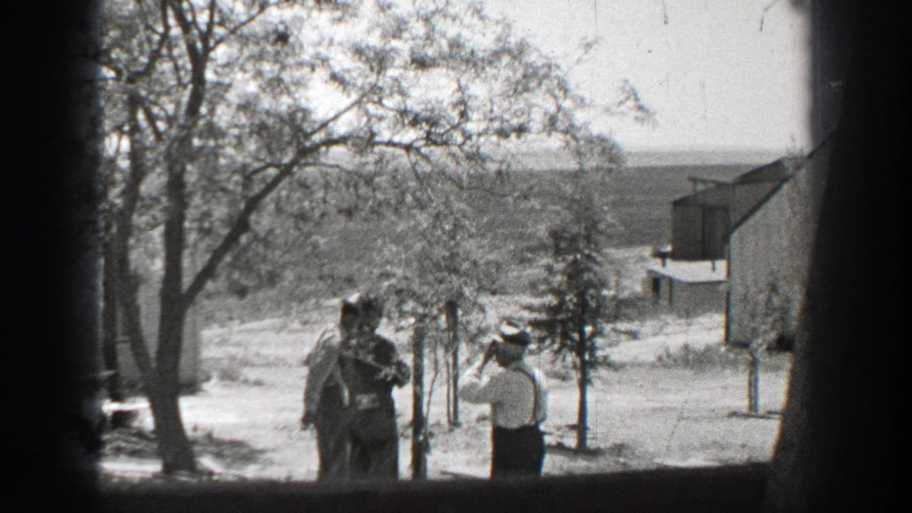 Three men stand in a sunny yard near young trees and simple buildings.