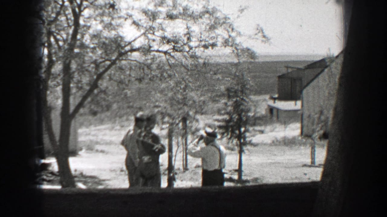 1937 California home-movie frame with a white-shirted man holding up an object toward two men in a sunny yard.