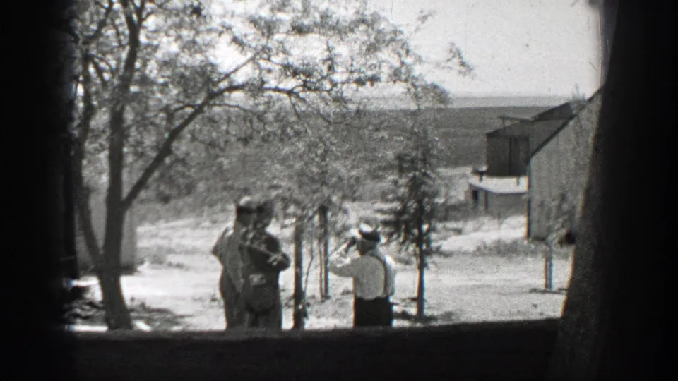 1937 California home-movie frame with a white-shirted man holding up an object toward two men in a sunny yard.