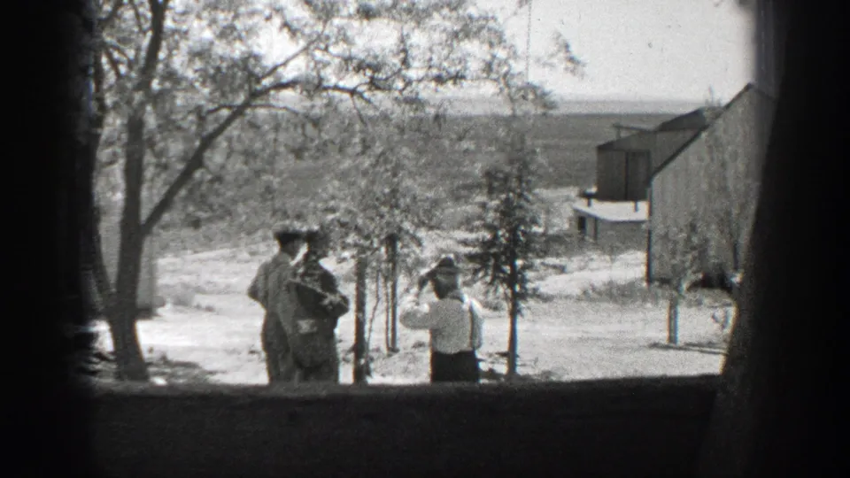 1937 California home-movie frame of a white-shirted man facing two men, one in uniform-like clothing, near trees and a building.