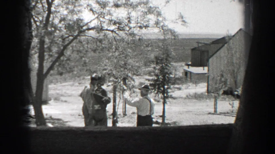 In 1937 California home-movie footage, two men in hats stand near young trees in a yard viewed through a window frame.