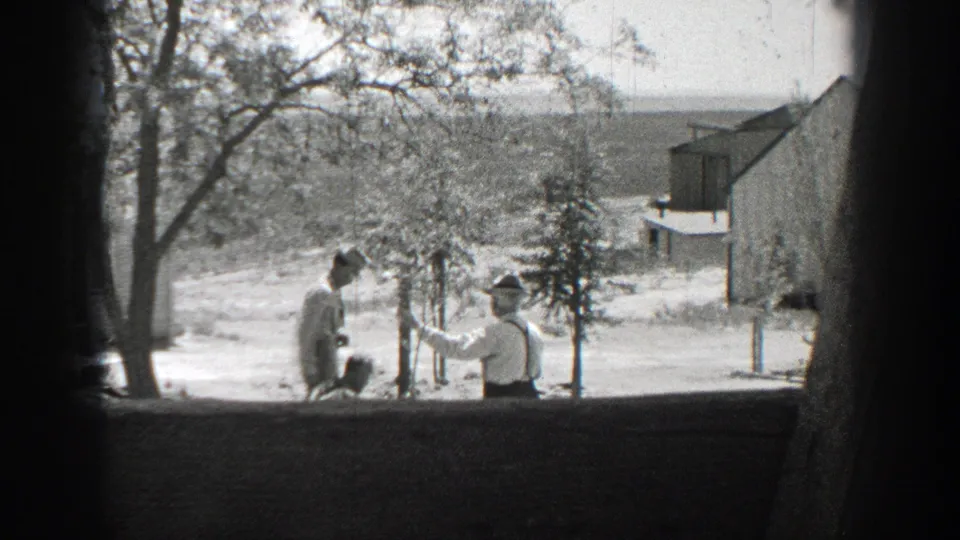 The yard scene continues as a woman pauses near a low figure and a man in a hat stands beside a young tree.