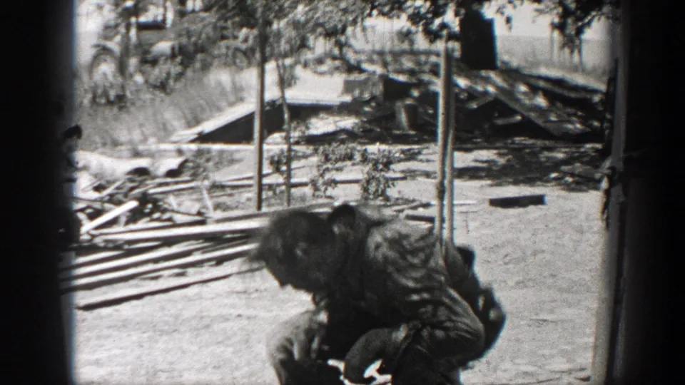 A man in work clothes bends near stacked wooden planks and scattered debris in a 1937 California home-movie yard scene.
