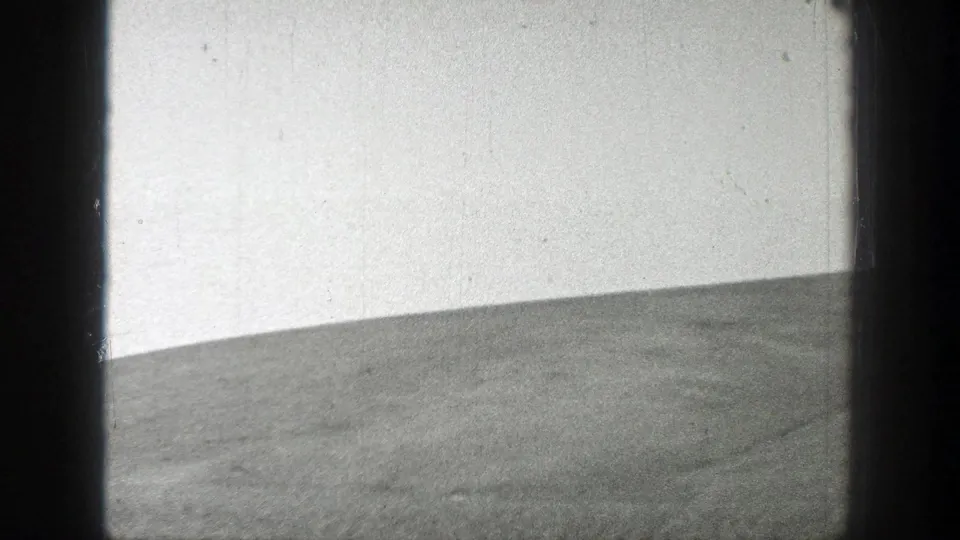 Black-and-white 8mm frame showing a low empty grassy hill in California, with pale sky, grain, and light scratches.