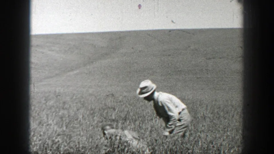1937 California 8mm frame of a man in a light shirt and brimmed hat bending in a grassy field.