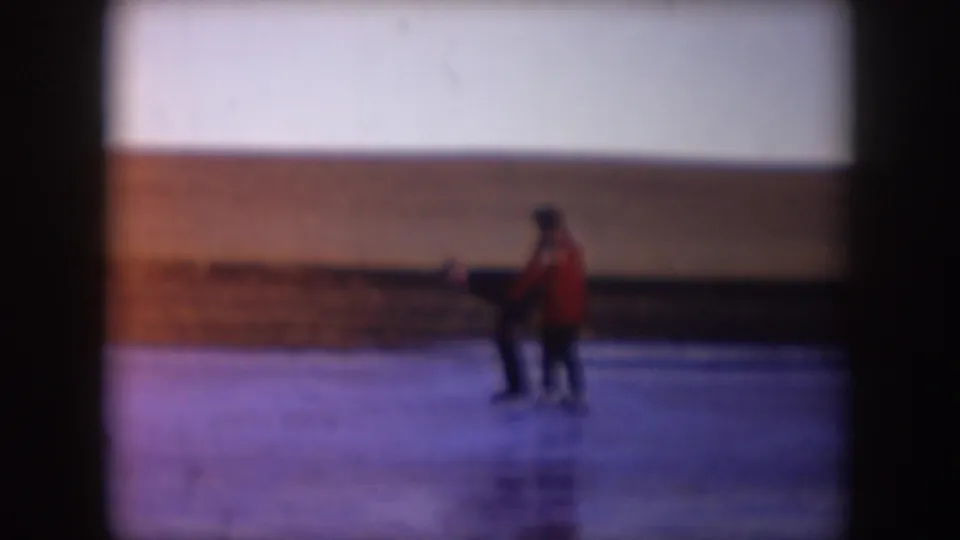 Two adults pause on the frozen lake, one in a red coat, with barren winter ground behind them.