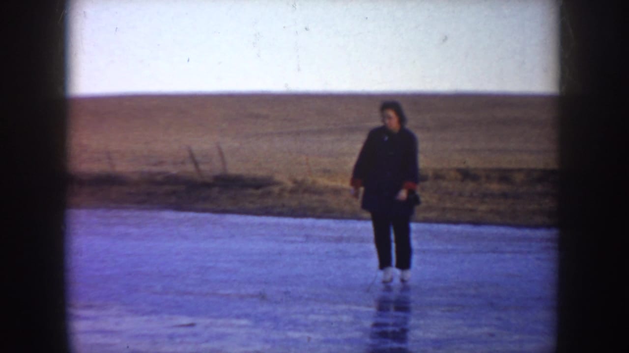 Woman in dark clothing stands on skates on a frozen pond in North Dakota, with a low open hill in the background.