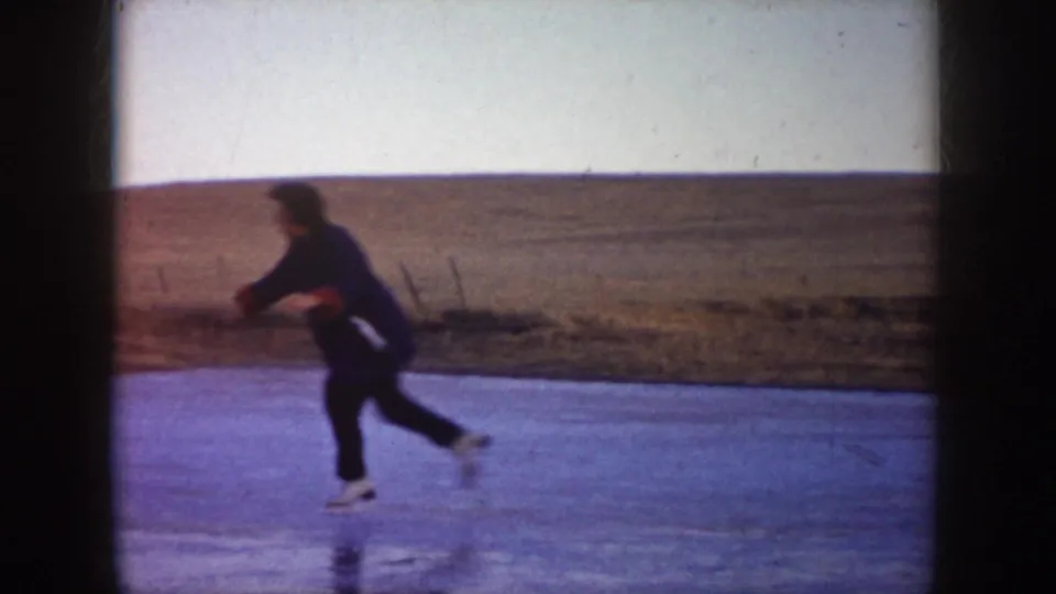 A woman skates across a frozen pond in 1950s North Dakota, moving in front of a broad, low hill.