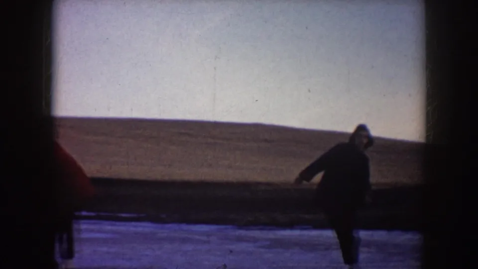 A woman in dark winter clothing skates near the right edge of a frozen North Dakota pond with open land behind her.