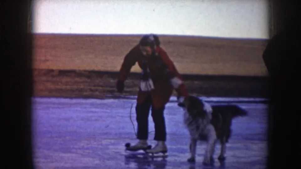 A red-coated skater moves beside a dog on a frozen lake in supported 1953 North Dakota 8mm home-movie footage.