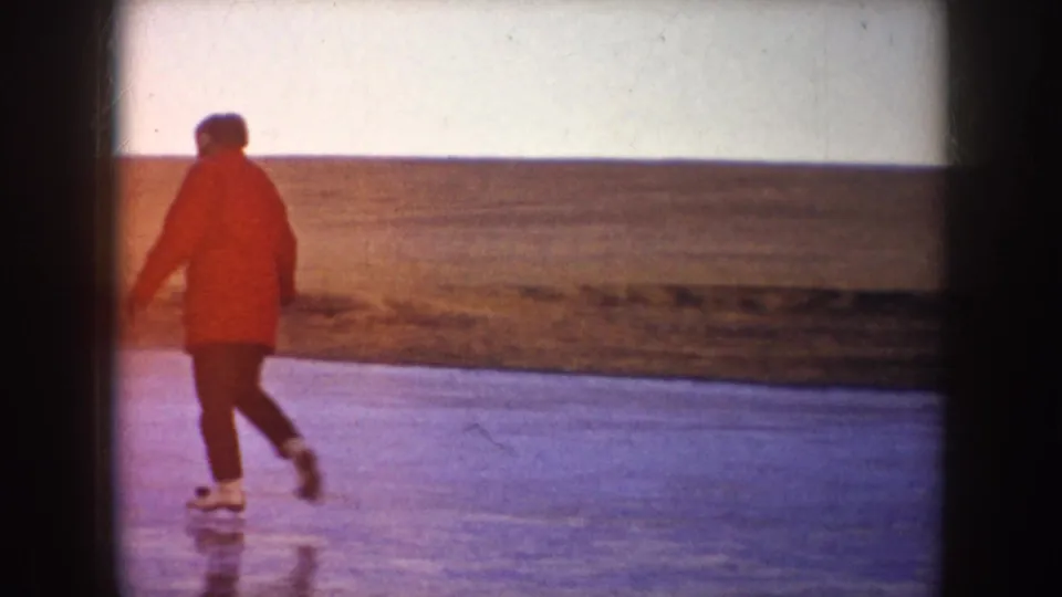A skater in a red coat crosses a frozen pond in the supplied 1953 North Dakota home-movie footage.