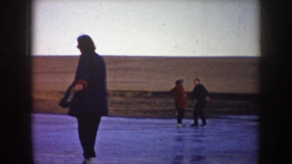 A bundled skater crosses the foreground as two distant skaters appear to hold hands on a North Dakota frozen pond.