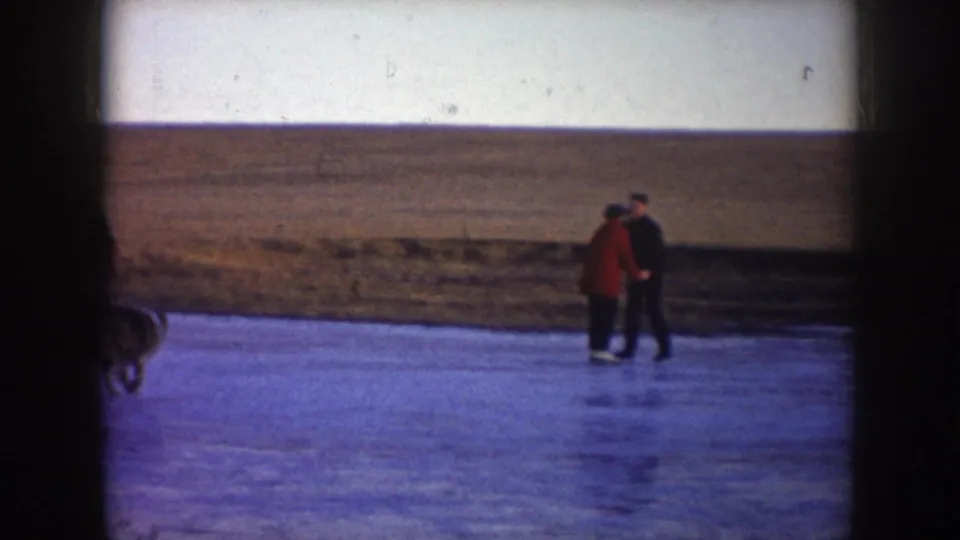 Two skaters, one in a red coat, hold hands on the ice in a 1953 North Dakota winter home-movie frame.