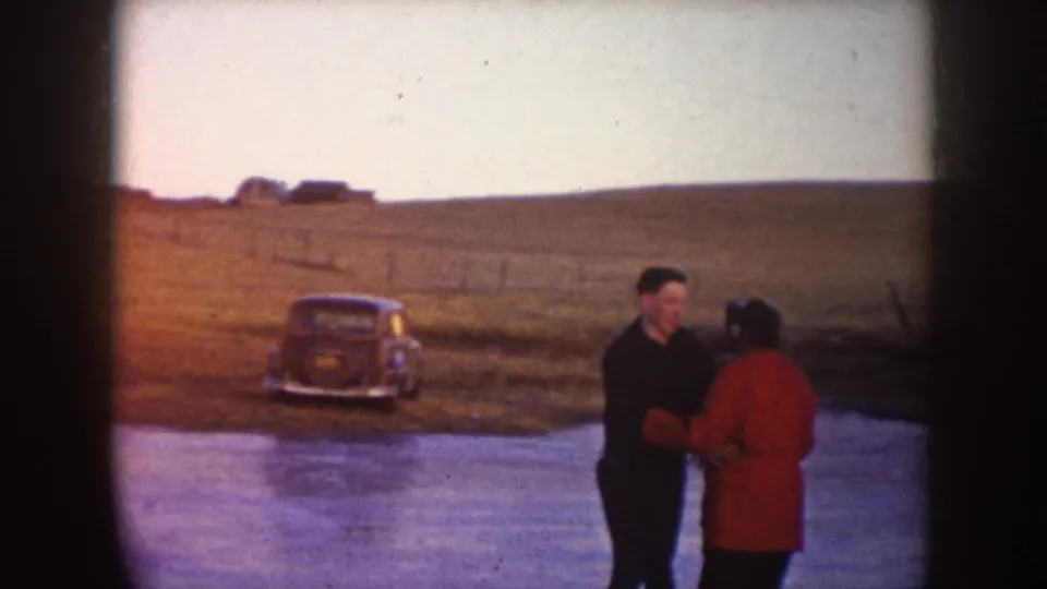 A couple appears to skate or steady themselves on a frozen pond in North Dakota, with a mid-century car parked on the grassy bank.