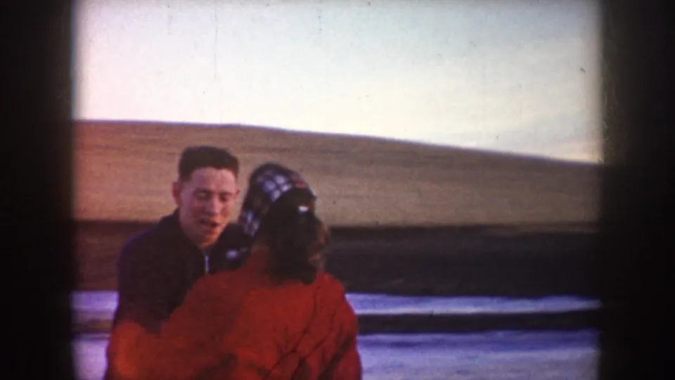 A closer 8mm frame shows the man facing camera while skating with the woman in a red coat on the frozen pond.