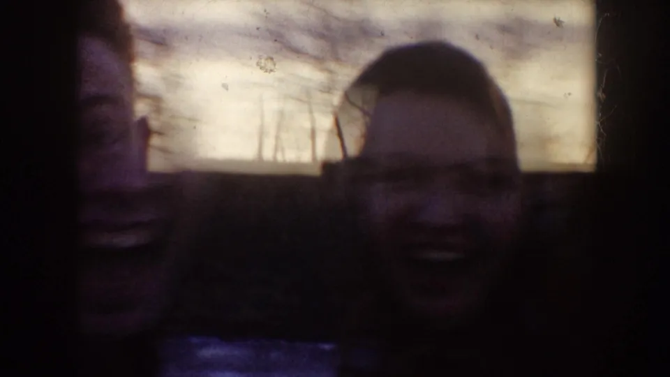 A soft, underexposed 1953 home-movie frame shows two faces close to the camera with winter landscape beyond.