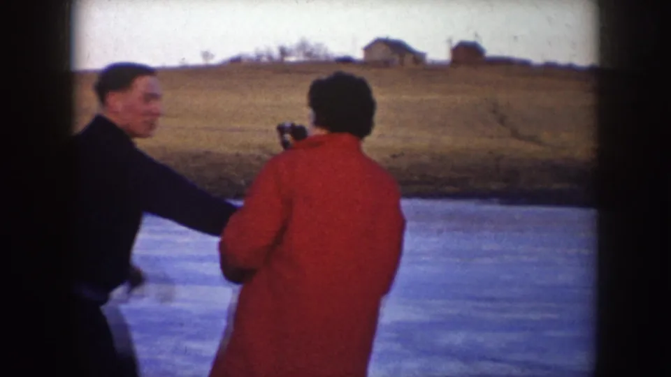 A man in a dark jacket reaches toward a woman in a red coat while they stand on ice in rural North Dakota, 1953.