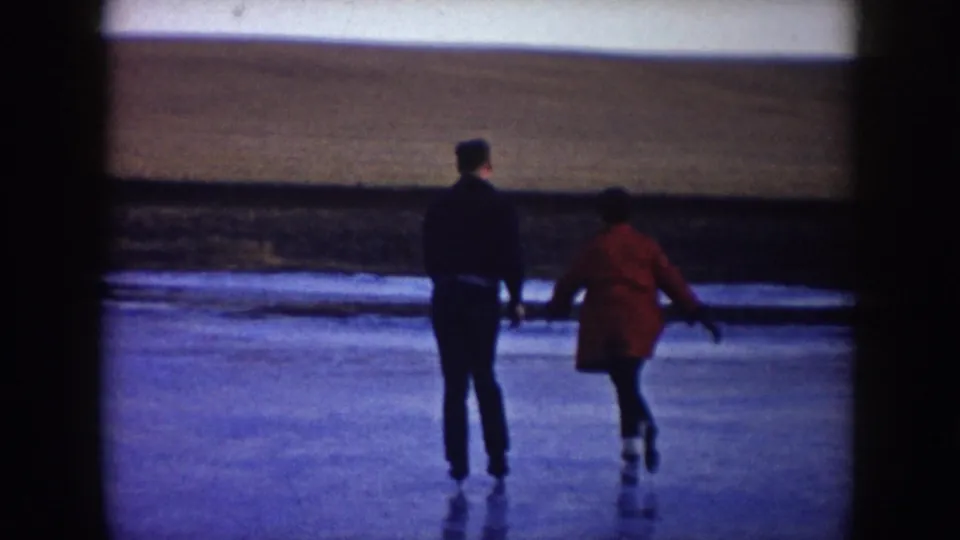 A 1953 North Dakota home-movie frame shows a man and woman skating away together across ice under an open winter sky.
