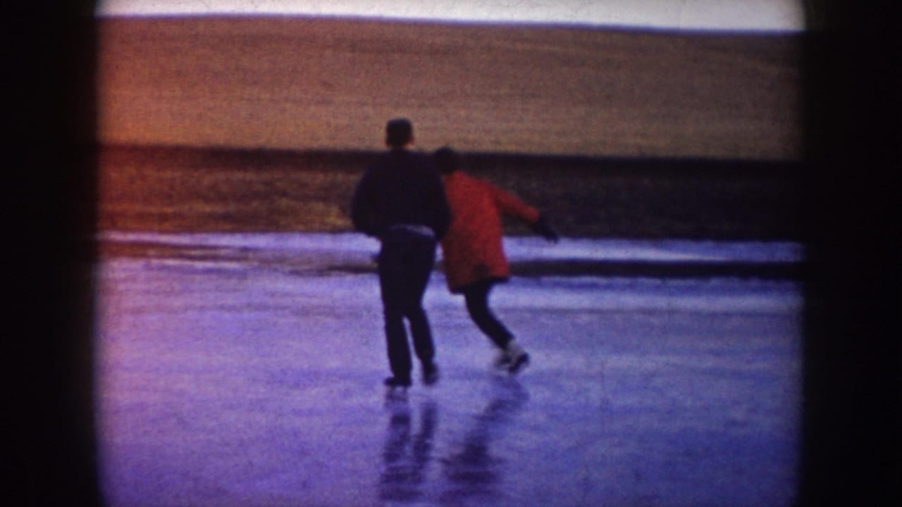 In 1953 North Dakota home-movie footage, two adults skate together on a frozen pond with open winter fields beyond.