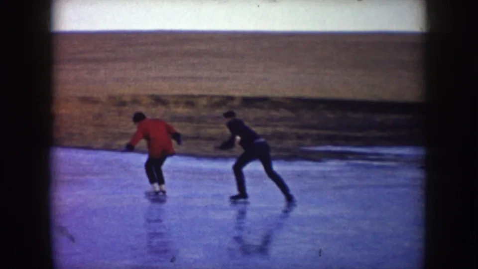 A red-coated skater leads across a North Dakota frozen pond while a second adult follows in dark clothing.