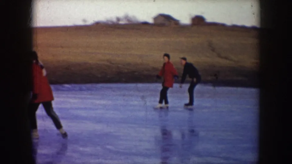 On the frozen pond, two adults skate past open North Dakota fields as a red-coated figure is partly cropped at the left edge.