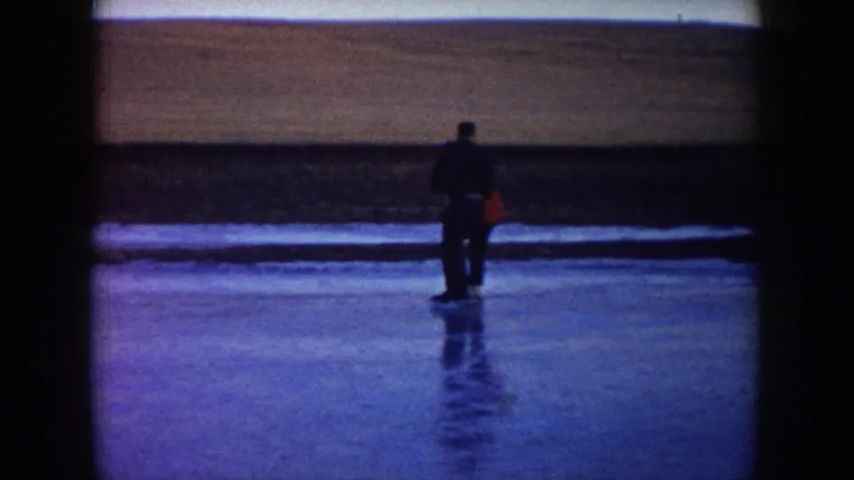 1953 North Dakota 8mm frame showing one adult skater in dark clothing on open ice near a low bank.