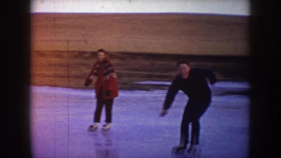 1953 North Dakota 8mm frame showing a woman in a red coat and a man in dark clothing skating on outdoor ice.