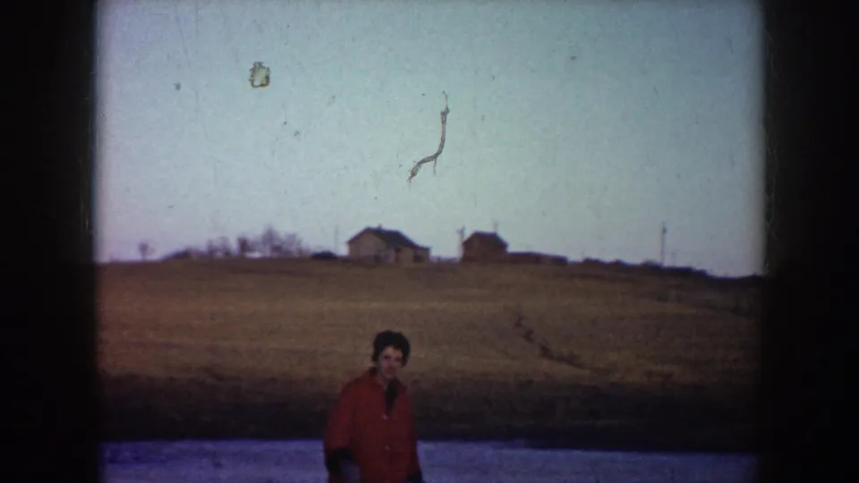 In 1953 North Dakota footage, a woman in a red coat is seen on a frozen pond with open ground and distant houses in the background.