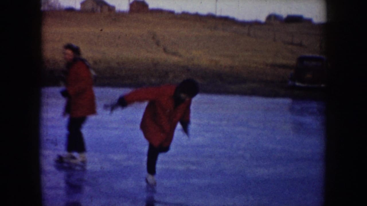 Two skaters move across a frozen pond in North Dakota, with a red coat prominent and an older parked car near the bank.