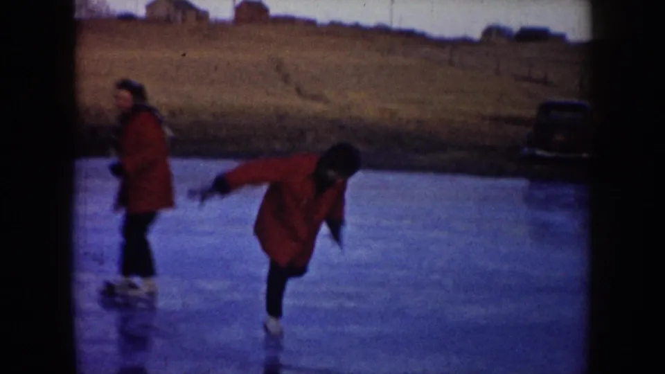 Two skaters move across a frozen pond in North Dakota, with a red coat prominent and an older parked car near the bank.