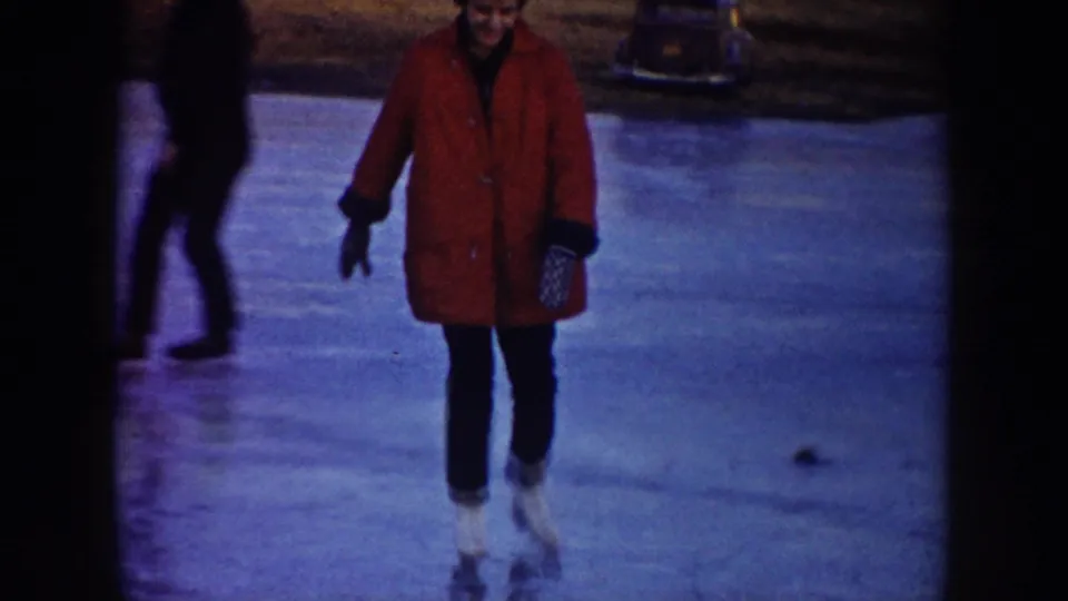 A woman in a red coat skates toward the camera on a North Dakota frozen pond as another person in dark clothing passes behind.