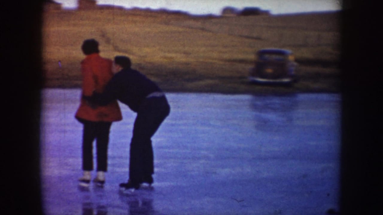 A dark-clothed skater appears to help a red-coated skater regain balance on the ice, with a parked older car near the bank.