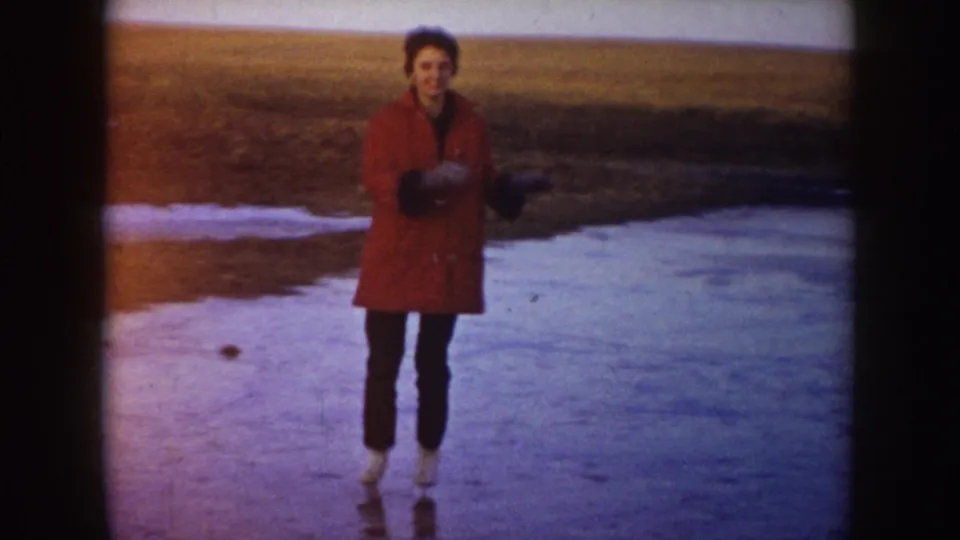 A woman in a red coat and dark pants faces the camera while ice skating on a frozen pond in North Dakota, 1953.