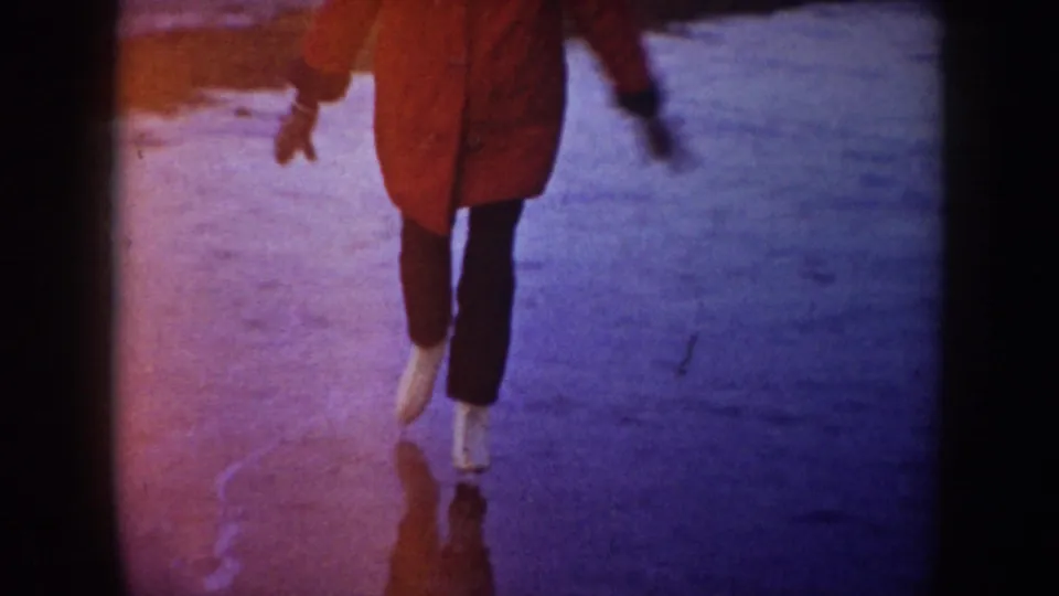 Close framing shows the skater's red coat, dark pants, and white skates moving across blue-toned pond ice.
