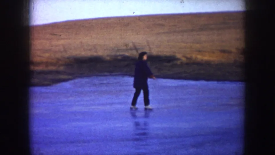 A skater moves across the frozen pond in a wide North Dakota home-movie view, with brown grassland beyond the ice.