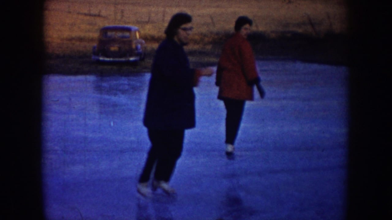 Two adults skate on outdoor ice in North Dakota, with a rounded 1950s car parked near the field behind them.