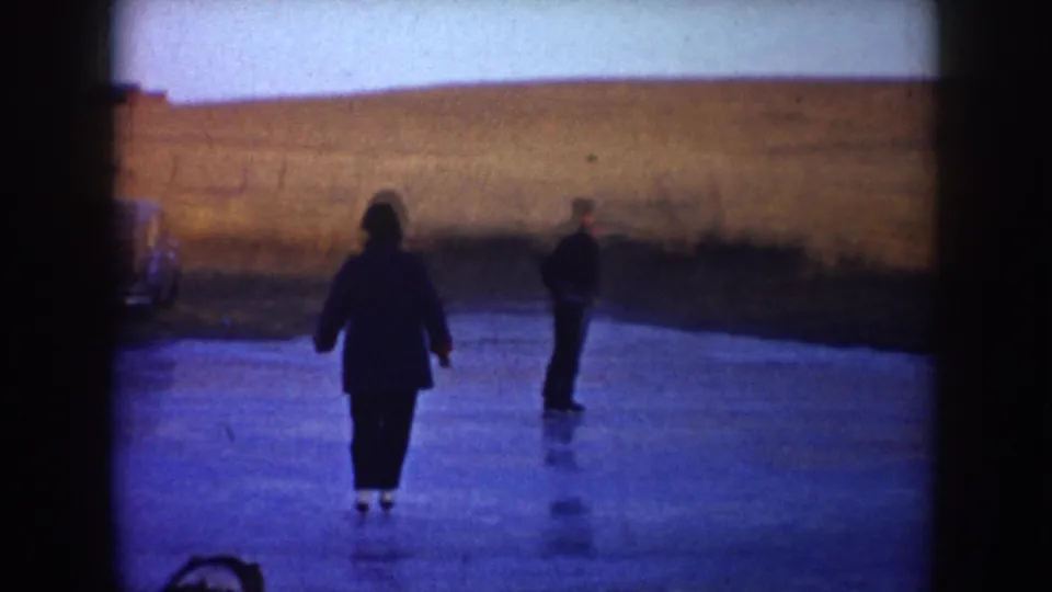 Two adult skaters on a frozen rural pond in North Dakota, seen in soft 1953 8mm home-movie color.