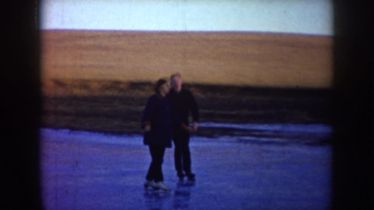 Two adult skaters pause together on a frozen pond in rural North Dakota, with brown fields beyond the ice.