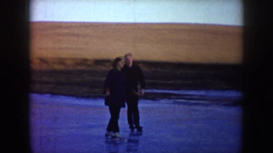 Two adult skaters pause together on a frozen pond in rural North Dakota, with brown fields beyond the ice.