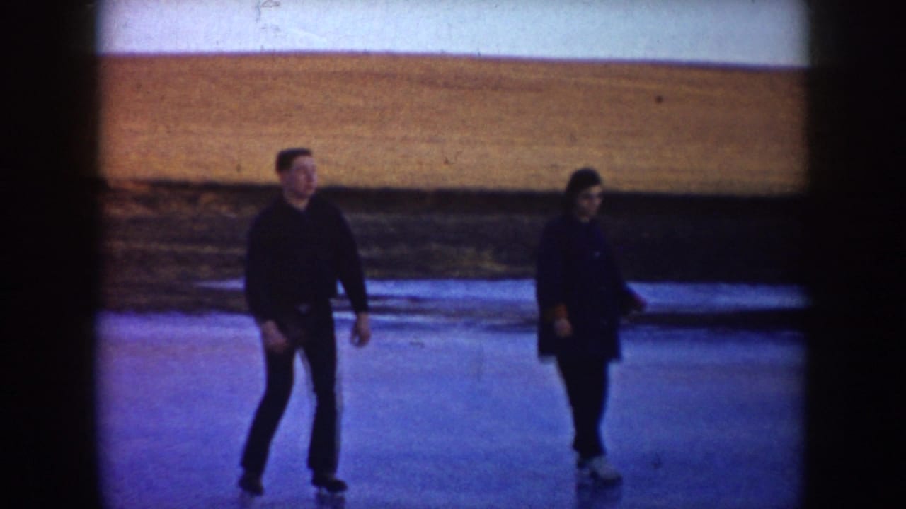 A man and woman skate on a frozen rural pond in North Dakota, framed by open fields and faded 8mm color.