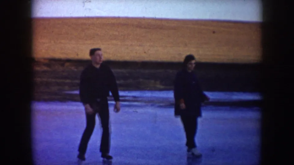 A man and woman skate on a frozen rural pond in North Dakota, framed by open fields and faded 8mm color.